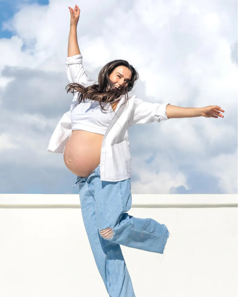 Happy pregnant woman jumping under blue sky, celebrating healthy and active motherhood lifestyle