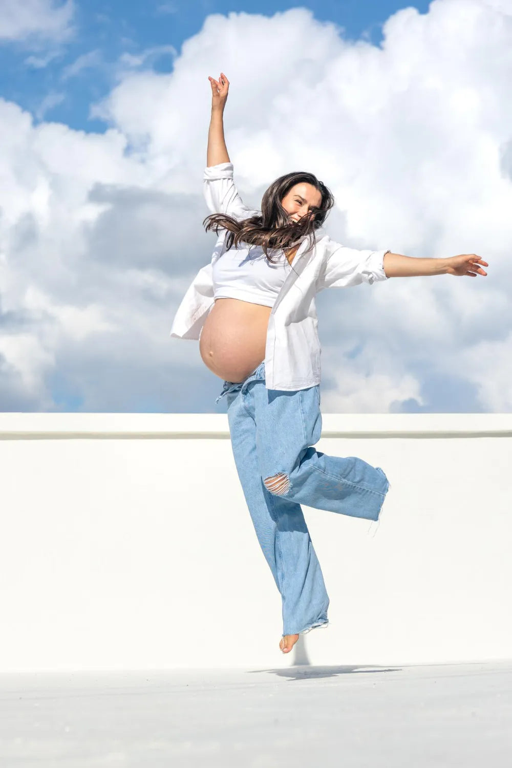 Happy pregnant woman jumping outdoors under blue sky, celebrating healthy and active motherhood lifestyle