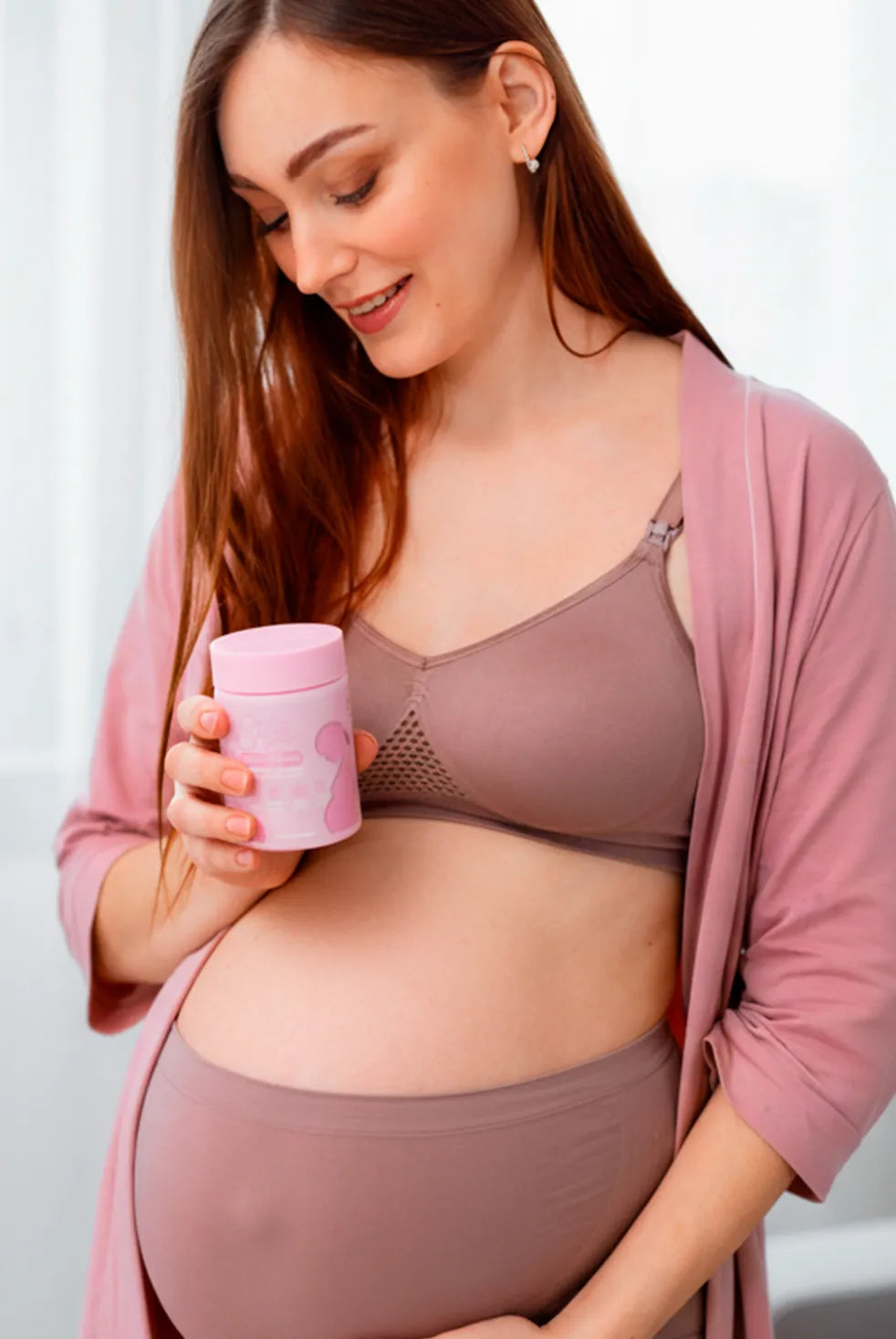 Pregnant woman holding pink prenatal vitamins supplement jar at home, maternity wellness lifestyle image